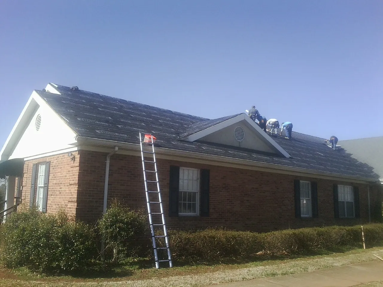 Crew tearing shingles off a brick ranch roof with a ladder set — Emergency Roof Repair work by our crew in Islip