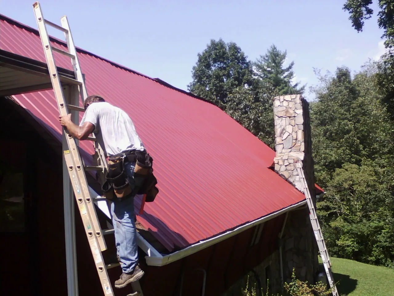 Roofer on a ladder beside a red metal roof and stone chimney — Soffit & Fascia Repair by Roofing Brothers in Islip